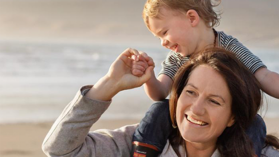 Mother carrying her child, a toddler, on her shoulders and smiling, walking on the beach